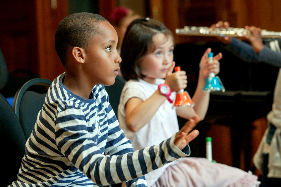 A young black boy, interacting with a musical activity, with a young girl, holding a pair of bells in their hands.